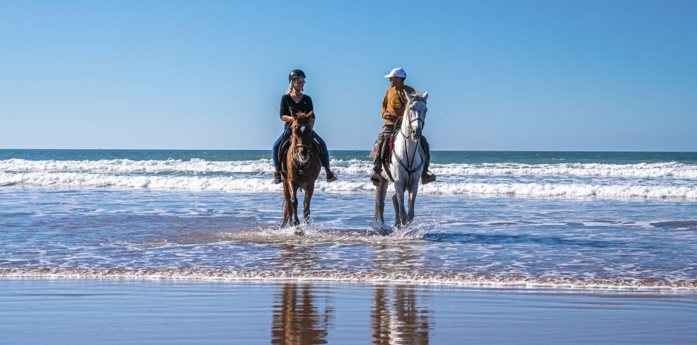 Man and woman riding horses along shoreline at beach against clear sky